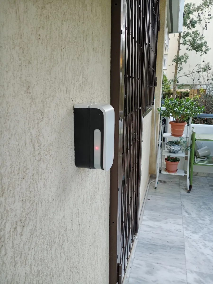 Εξωτερικός ανιχνευτής με αναμμένο κόκκινο LED, στον τοίχο δίπλα σε πτυσσόμενο κάγκελο μπαλκονιού.